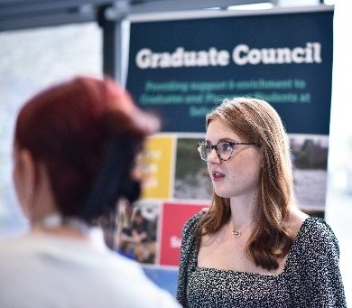 A graduate in front of a Graduate Council display banner